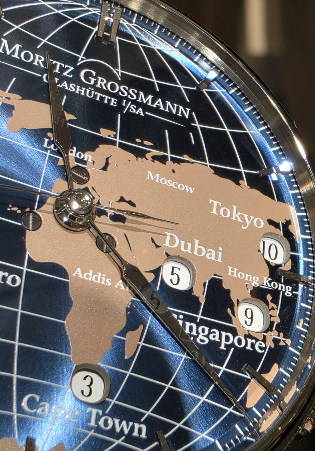 Close-up of a Moritz Grossmann world map clock dial, showing continents in beige over a blue globe background with white grid lines and city names. OsterJewelers.com