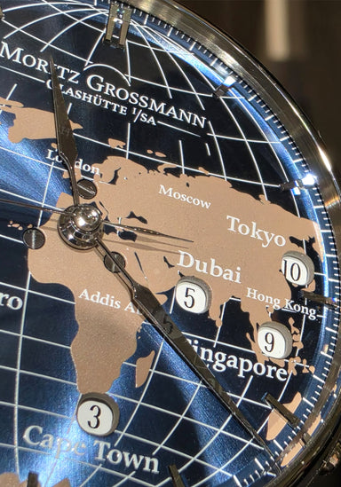Close-up of a Moritz Grossmann world map clock dial, showing continents in beige over a blue globe background with white grid lines and city names. OsterJewelers.com
