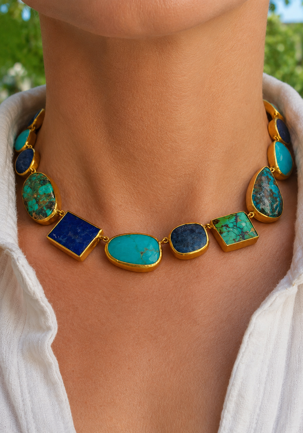 A close-up of amodel wearing a yellow gold-statement multi colored turquoise necklace featuring multiple turquoise, lapis lazuli, and green stone cabochons, paired with a white collared shirt. Oster Jewelers