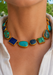 A close-up of amodel wearing a yellow gold-statement multi colored turquoise necklace featuring multiple turquoise, lapis lazuli, and green stone cabochons, paired with a white collared shirt. Oster Jewelers
