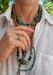 A close-up of a model wearing a 18kyg  multi colored turquoise necklace featuring multiple turquoise, lapis lazuli, and green stone cabochons, paired with a white collared shirt. Paired with layering necklaces from Oster Jewelers