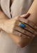 A close-up of a hand wearing a yellow gold ring with a teardrop-shaped blue labradorite stone, set against a soft beige dress.