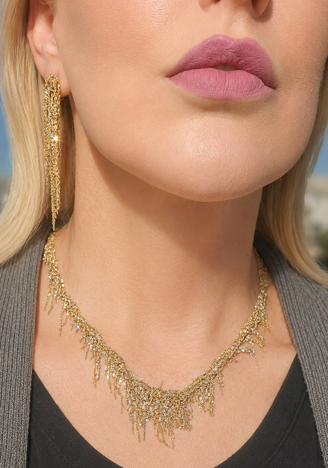 Close-up of a model wearing a gold-toned fringed necklace and matching long drop earrings, with visible skin, lips, and a gray top against a softly blurred outdoor background.