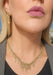 Close-up of a model wearing a gold-toned fringed necklace and matching long drop earrings, with visible skin, lips, and a gray top against a softly blurred outdoor background.