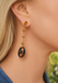 A close-up of model wearing a dangle earring made from ebony. Earring features a gold-toned post with a round amber top, several small gold disces, and a large black oval drop with cutouts and tiny gold and red accents.